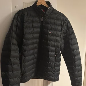 Svart pufferjacka från Tommy Hilfiger - Säljer en svart pufferjacka från Tommy Hilfiger. Jackan har en stilren design med dragkedja och långa ärmar. Perfekt för kyligare dagar med sin quiltade look. Den har en liten logga på bröstet och ribbade detaljer vid kragen.