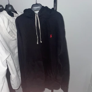 Svart hoodie från Polo Ralph Lauren - Säljer en klassisk svart hoodie från Ralph Lauren med det ikoniska röda logotypen på bröstet. Hoodien har en justerbar dragsko i huvan och en stor ficka framtill. Perfekt för en avslappnad stil. Skulle säga den passar folk runt 165.