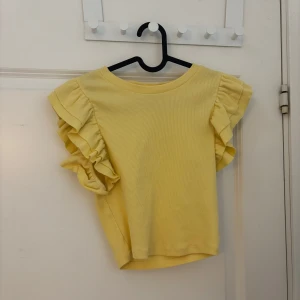 Gul ribbad topp från Zara - Säljer en söt gul ribbad topp från Zara med volangdetaljer på ärmarna. Toppen har en rund halsringning och korta ärmar. Perfekt för en solig dag!