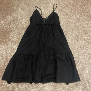 Svart, kort klänning från Zara - Snygg svart klänning från Zara med smala axelband och v-ringning. Klänningen har en volangdetalj nedtill och är perfekt för en stilren look. Tillverkad i ett lätt och bekvämt material. (Justerbar med band 150 kr vid snabbköp)