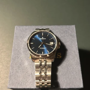 Elegant klocka från Morris - Snygg Morris-klocka med en stilren blå urtavla och datumvisning. Boetten och armbandet är i silverfärgat rostfritt stål. Perfekt för den som vill ha en tidlös accessoar med en modern touch. Kommer i originalförpackning. Original pris 2900