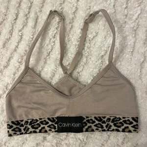 Beige linnetopp från Calvin Klein - Säljer en snygg beige sport bh från Calvin Klein med leopardmönstrad kant. Toppen har justerbara axelband och är perfekt för en stilren look. Den är tillverkad i ett mjukt material som ger en bekväm passform.