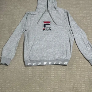 Grå hoodie från FILA - Säljer en stilren grå hoodie från FILA med deras ikoniska logga i rött och svart på bröstet. Tröjan har en justerbar huva med dragsko och en praktisk magficka. Perfekt för en avslappnad stil.