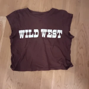Brun t-shirt med tryck 'Wild West' - Säljer en brun t-shirt med texten 'Wild West' i vitt på framsidan. T-shirten har en avslappnad passform och är ärmlös, perfekt för en casual look.
