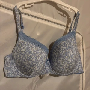 Blå blommig bh med spetsdetaljer - Söt blå bh med vita blommor och spetsdetaljer. Den har en liten rosett framtill och justerbara axelband för perfekt passform. Ej använd.