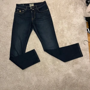 Mörkblå jeans från Morris - 29 32Snygga mörkblå jeans från Morris med klassisk femficksdesign och guldfärgade detaljer. Jeansen har en normal passform och är tillverkade i ett slitstarkt denimtyg. Perfekta för en stilren look.