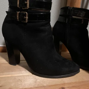Svarta stövletter med spännen stl 40  - Snygga svarta stövletter med klack och dekorativa spännen. Skorna har en dragkedja på sidan för enkel på- och avtagning. Perfekta för att ge en stilren touch till din outfit.