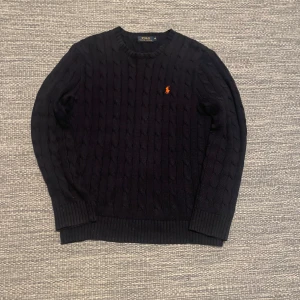 Marinblå kabel stickad tröja Polo Ralph Lauren - Säljer en stilren svart stickad tröja från Polo Ralph Lauren med det klassiska logotypbroderiet i orange på bröstet. Tröjan har en rund halsringning och långa ärmar, perfekt för kyligare dagar. Mycket bra skick. Ca 2000 kr mitt pris 649 kr