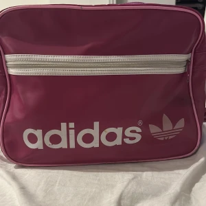 Rosa axelväska från Adidas - Säljer en snygg rosa axelväska från Adidas lite av den vintage stuket med vit dragkedja och logga. Väskan har ett stort huvudfack och en praktisk ytterficka med dragkedja. Bra plats i väskan med me 2 innerfickor