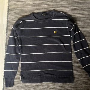Randig tröja från Lyle & Scott - Säljer en snygg randig tröja från Lyle & Scott i mörkblått med vita ränder. Tröjan har ett gult logomärke på bröstet och är långärmad med ribbade muddar. Perfekt för en stilren look!