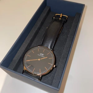 Daniel Wellington klocka - En Daniel Wellington klocka med svart läderband samt svart urtavla. Visarna samt resten av detaljerna är i en rose-guld färg. Klockan är i bra skick och köptes ny för ~1200kr. Lådan samt ”instruktionsbok” medföljer. 