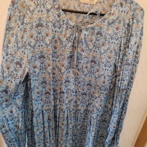 Blommig tunika I siden från Odd Molly, stl 2 - Vackert blå tunika från Odd Molly  i siden med ett vackert blommönster i beige och mörkblå toner. Tunikan har långa ärmar med och en knytning framtill vid halsen.hel och ren, inga djur i hemmet.
