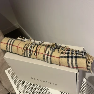 Designerhalsduk Burberry inspirerad  - Stilren beige halsduk med klassiskt rutigt mönster i svart och rött. Passar både män och kvinnor. Perfekt för att hålla sig varm och stilren under kyliga dagar. Den har fransar i ändarna som ger en extra touch av elegans.