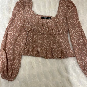 Beige blus från Missguided - Säljer en söt beige blus från Missguided med ett vitt abstrakt mönster. Blusen har långa ärmar och en smockad midja som ger en fin passform. Perfekt för en stilren look.