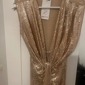 Glittrig guldklänning från Lisa Style - Snygg glittrig guldklänning från Lisa Style. Klänningen har en djup v-ringning och är täckt med paljetter som ger en glamorös look. Perfekt för att sticka ut på festen! ✨