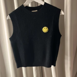 Svart stickad väst med smiley - Snygg svart stickad väst med en gul smiley på bröstet. Perfekt för att lägga till en touch av humor och stil till din outfit. Västen har en ribbad struktur och är ärmlös, vilket gör den lätt att matcha med olika plagg.