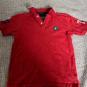 Röd pikétröja från Sail Racing - Snygg röd pikétröja från Sail Racing med broderad detalj på ärmen och ett märke på bröstet. Tröjan har en klassisk krage med knappar och är perfekt för en sportig look. Storlek 140.
