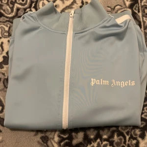 Ljusblå track jacket från Palm Angels - Säljer en snygg ljusblå track jacket från Palm Angels med vit dragkedja och logga på bröstet. Jackan har långa ärmar och ribbade muddar. Perfekt för en stilren look.