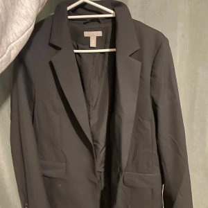 Svart kavaj från H&M - Snygg svart kavaj från H&M med klassisk design. Kavajen har en stilren passform och är försedd med knappar vid ärmen. 