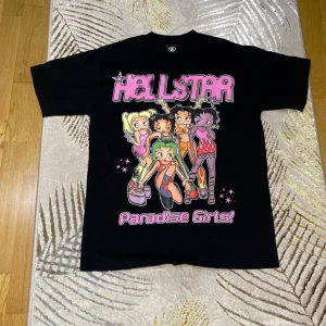 Svart t-shirt med Paradise Girls-tryck - Cool svart t-shirt med färgglatt tryck av Paradise Girls på framsidan och en stiliserad profil med flammor på baksidan. Tillverkad i 100% bomull för en bekväm känsla. Perfekt för dig som gillar unika och uttrycksfulla plagg.