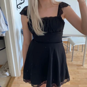 Svart klänning med spetsdetaljer - Snygg svart klänning med spetsdetaljer på axelbanden och längs kanten. Klänningen har en öppen rygg med snörning. Bara provad men hade en defekt vid midjan som med lösa trådar. Men går lätt att fixa med symaskin.  Den är utformad lite annorlunda så man knyter den i midjan med ett löstagbart snöre.