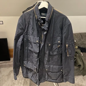 jacka från Polo Ralph Lauren - Säljer en snygg blå jacka från Polo Ralph Lauren. Jackan har flera fickor med knappar och dragkedja, samt en hög krage. Perfekt för en stilren look.