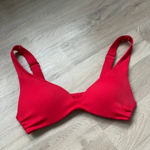 Röd ribbad bikiniöverdel från H&M - Snygg röd bikiniöverdel från H&M med ribbad textur. Den har breda axelband och en praktisk knäppning bak. Perfekt för en dag på stranden eller vid poolen. 🔥bikinin är hel men lite tråd har gått upp (se bilder). 