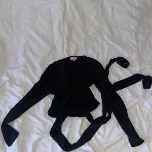 Pilates, omlott tröja/topp - Snygg svart omlottopp från L'Academie i ett mjukt och glansigt material. Toppen har långa ärmar och ett knytband i midjan för en justerbar look. Passar L