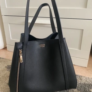 Svart axelväska från Guess - Snygg svart axelväska från Guess i mycket bra skick. Väskan har en stilren design med dragkedjedetalj och är perfekt för både vardag och fest. Tillverkad i syntetmaterial med en elegant finish.