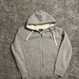 Polo Ralph lauren zip - Säljer en snygg grå hoodie från Polo Ralph Lauren. Den har en dragkedja framtill och en broderad logga på bröstet. Perfekt för höst och vinter med sin mjuka insida. Storleken är xl men passar deffenetift som L
