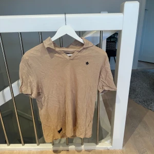 Beige kortärmad t-shirt - Säljer en beige kortärmad t-shirt i bomull. Den har en liten broderad detalj på bröstet och en avslappnad passform. Perfekt för sommardagar eller som ett lager under en jacka.