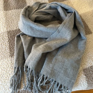 Grå halsduk - Säljer en supermjuk och stilren grå halsduk med fransar i ändarna. Perfekt för att hålla sig varm under kyliga dagar. Den är i nyskick och bra nu för vintern. Enkelt att matcha med olika outfits!