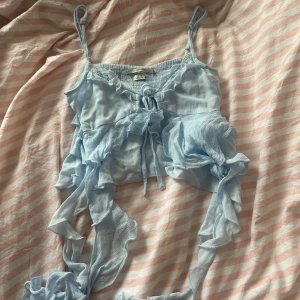 Ljusblå volanglinnetopp - Säljer en superfin ljusblå linnetopp med volanger och knytning framtill. Toppen är ärmlös och har smala axelband. Den är croppad och ger en söt och luftig look. Passar perfekt till jeans eller kjol!