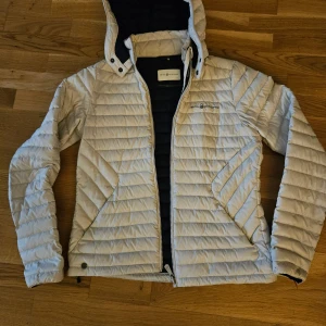 Vit quiltad jacka från Sail Racing - Säljer en snygg vit quiltad jacka från Sail Racing. Den har en dragkedja framtill och en huva med svarta detaljer. Perfekt för kyliga dagar och har en stilren design. Jackan är i mycket bra skick och passar perfekt för höst och vår. Använd endast en enda gång. 