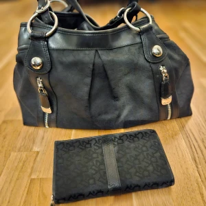 Svart väska och plånbok från DKNY - Säljer en snygg svart väska och matchande plånbok från DKNY. Väskan har silverfärgade detaljer och dragkedjor, perfekt för både vardag och fest. Plånboken har flera fack och en praktisk dragkedja. Båda är i mycket bra skick och har ett diskret mönster som ger en lyxig känsla. Utan skavanker på utsidan, fläckar på insidan. Du får plats med datorn i den. 