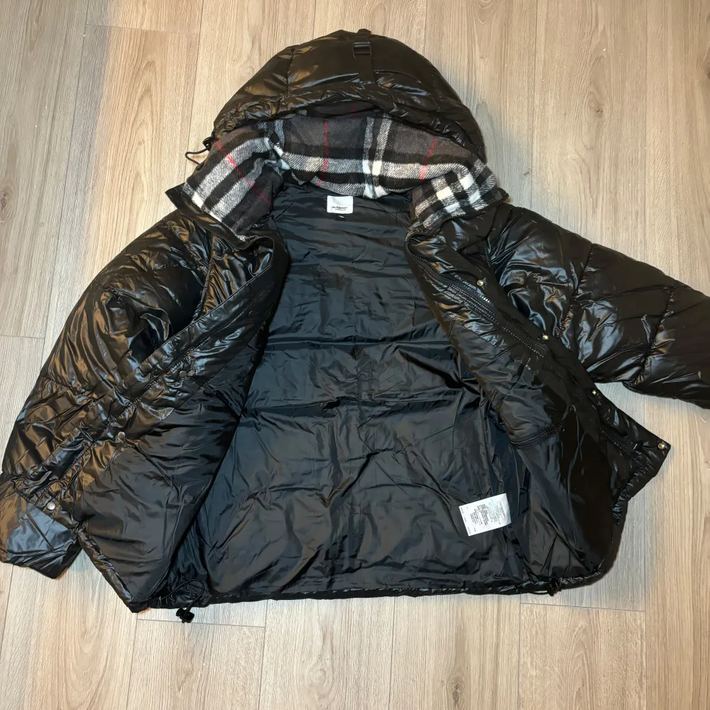 Burberry Pufferjacka Snygg och elegant svart pufferjacka från Burberry, perfekt för kyliga vinterdagar. Jackan har en glänsande, quiltad yta och en klassisk huva fodrad med Burberrys ikoniska rutmönster. . Jackor.