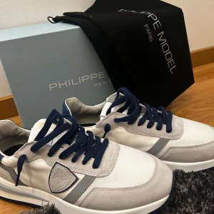 Philippe Model sneakers i vitt och blått - Snygga Philippe Model sneakers i vitt och blått med en stilren design. Skorna har en vit bas med blå detaljer och mörkblå snörning. De är i nyskick och perfekta för både vardag och fest. Sulan är robust och ger bra grepp. Kommer med originalförpackning. Säljer dom då dom inte passade mig  ( priset är inte skrivet i Sten)