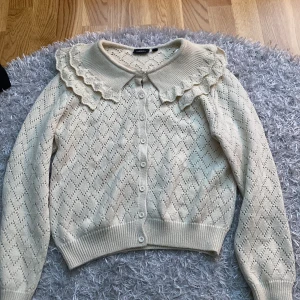 Beige stickad kofta med volangkrage - Superfin beige stickad kofta med volangdetaljer runt kragen. Den har ett diskret mönster och knappar hela vägen fram. Perfekt för höst och vår när du vill vara både stilig och varm. Passar till både jeans och kjol!