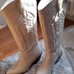Beiga cowboy boots - Snygga vita cowboy boots med dekorativa sömmar och högt skaft. De har en låg klack och spetsig tå. Dom är använda ett fåtal gånger då dom va för små för mig med det var för sent att lämna tillbaka. Inga synliga skador. Skriv för fler bilder.