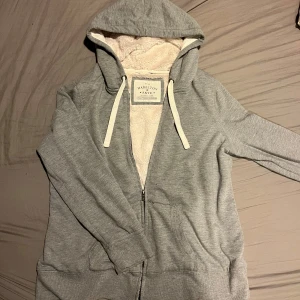 Fodrad kofta - Mysig grå hoodie med teddyfoder Den har en dragkedja framtill och en stor huva med vita snören. Perfekt för kyliga dagar när du vill hålla dig varm och bekväm.