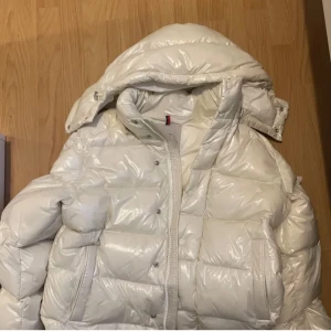 Vit dunjacka från Moncler - Säljer en snygg vit dunjacka från Moncler med glansig finish. Jackan har en hög krage och en avtagbar huva. Den är försedd med dragkedja och tryckknappar framtill samt Moncler-logga på ärmen. Perfekt för kyliga dagar! Använd väl och med kärlek så inga defekter, använd denna vinter. Säljer för att jag vill köpa en vår jacka🔥 Inget kvitto ingår tyvärr. Farsan köpte den i julklapp från plick så kan inte lova att den är äkta men väljer att tro på farsans ord💯