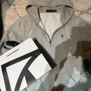 Grå hoodie från Ralph Lauren - Säljer en stilren grå hoodie från Ralph Lauren med dragkedja och den klassiska loggan broderad i blått på bröstet. Perfekt för en avslappnad look med justerbar huva och bekväma fickor. Passar perfekt för kyliga dagar.