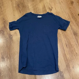 Mörkblå t-shirt från D.M.D by denim day - Säljer en stilren mörkblå t-shirt från D.M.D by denim day. T-shirten har en liten bröstficka och rund halsringning. Perfekt för en avslappnad look.