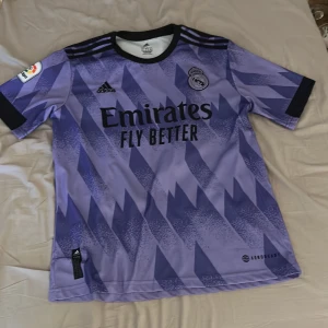 Real Madrid tröja - Säljer en snygg lila fotbollströja från Adidas med Real Madrid-logga och sponsortryck. Benzema på ryggen nummer 9. Tröjan har korta ärmar och ett coolt mönster i olika nyanser av lila. Perfekt för fotbollsfans! 🟣⚽