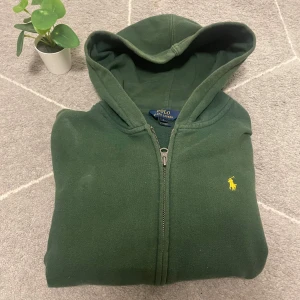 Grön hoodie från Ralph Lauren - Säljer en snygg grön hoodie från Ralph Lauren med dragkedja och huva. Den har en gul broderad logga på bröstet och praktiska fickor framtill. Perfekt för en avslappnad stil. Hör av dig vid intresse! 🌿