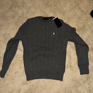 Grå stickad tröja från Ralph Lauren - Säljer en stilren grå stickad tröja från Ralph Lauren med det klassiska logotypbroderiet på bröstet. Tröjan har långa ärmar och ribbade muddar vid ärmslut och nederkant. Perfekt för kyligare dagar.