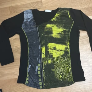 Svart och grön tröja från Micha - Unik svart tröja från Micha med gröna och gråa mönsterdetaljer. Tröjan har en dragkedja framtill och ett abstrakt mönster med zebraränder och text. Perfekt för dig som vill sticka ut med en kreativ stil.