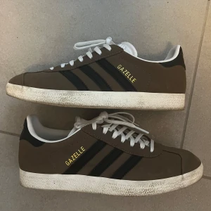Bruna Adidas Gazelle sneakers - Snygga bruna Adidas Gazelle sneakers med svarta ränder och vit sula. Skorna har klassisk snörning och är tillverkade i mocka. Perfekta för en stilren look. Använda få tillfällen, säljes på grund av fel storlek. 37 1/3 