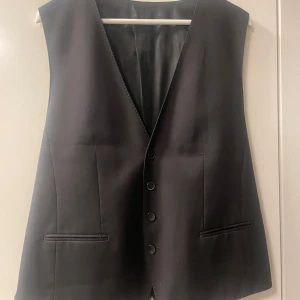 Svart väst från Hugo Boss - Elegant svart väst från Hugo Boss med knappar framtill och en justerbar rem på baksidan. Tillverkad i ett stilrent material från Guabello, perfekt för en sofistikerad look.