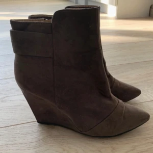 Bruna kilklackade boots - Snygga bruna boots med kilklack och spetsig tå. Perfekta för en stilren look. Tillverkade i mocka med dragkedja på sidan för enkel på- och avtagning.